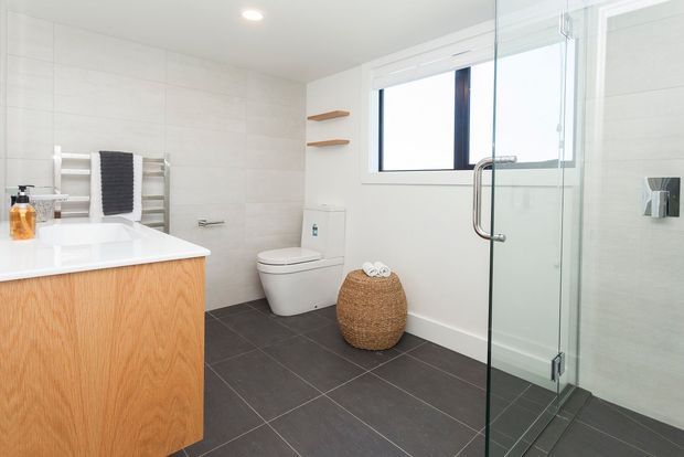 Bright contemporary bathroom with a floating timber vanity, white basin, wall-mounted toilet, dark large-format floor tiles, pale tiled walls, heated towel rail, and a frameless glass walk-in shower. A small woven basket and open timber shelves add warmth.