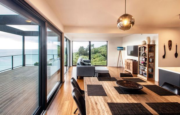 Open-plan contemporary living and dining area with expansive floor-to-ceiling glazing opening onto a timber deck and coastal view. The room features warm timber flooring, a large timber dining table with dark woven placemats, black dining chairs, a grey sofa, built-in media cabinetry, shelving, and a spherical pendant light. Dark-framed glazing and abundant natural light create a polished, airy interior.