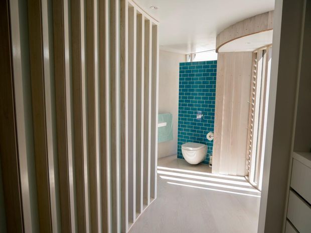 A bright contemporary bathroom viewed from the adjacent circulation space, featuring a wall-hung white toilet against a vivid turquoise-blue rectangular tiled feature wall, pale timber or wood-look screening and curved joinery, white walls, and strong linear sunlight filtering through vertical slats. A partial vanity or storage unit is visible at right.