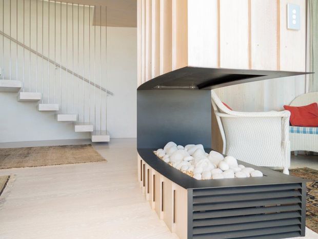 Bright contemporary interior featuring a sculptural black built-in fireplace with horizontal ventilation slats and a pebble-filled hearth. Pale timber wall panelling, light flooring, a floating staircase with slim vertical balustrade wires, and a partially visible lounge chair create a clean, refined setting.