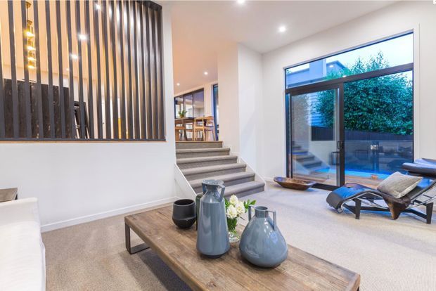 Bright contemporary open-plan living room with neutral carpet, a large timber coffee table, white sofa, and modern lounge chairs. A partially screened stairway with vertical timber slats leads to a raised dining area, while full-height glazing opens toward a small courtyard or pool area.