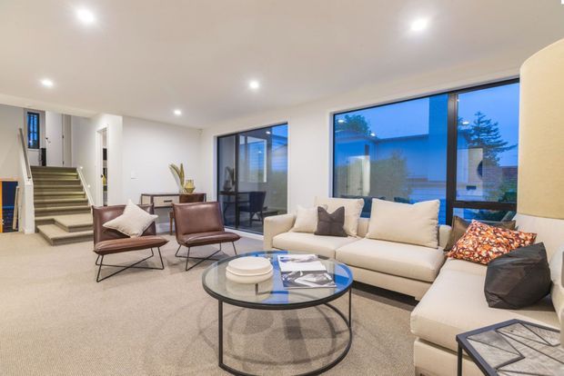 Bright contemporary living room with a large cream sectional sofa, brown leather lounge chairs, a round glass coffee table, recessed ceiling lights, and expansive dark-framed windows. A stairway and entry circulation area are visible in the background, with evening light outside.