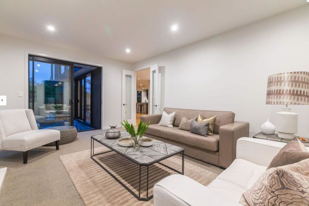 Bright contemporary living room with a neutral palette, upholstered sofas and armchair, a black-framed glass door opening to an outdoor area, recessed ceiling lights, a glass-topped metal coffee table, and a large textured rug. A hallway and adjoining spaces are visible beyond the room.
