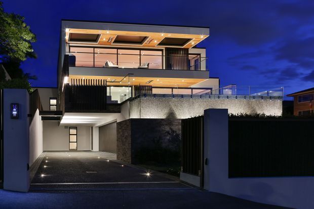 Night-time exterior view of a contemporary multi-level residence. The facade features broad glazed openings, projecting balcony terraces, warm recessed ceiling lighting, dark vertical screening, textured masonry walls, and a paved driveway leading to a recessed garage or entry area. The composition emphasizes the building envelope and dramatic architectural lighting.