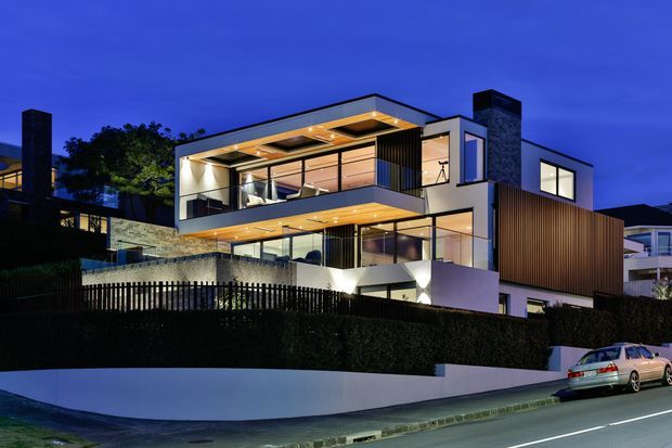 Twilight exterior view of a large multi-level contemporary residence on a sloping suburban site. The home features projecting flat-roofed volumes, extensive floor-to-ceiling glazing, illuminated timber-lined soffits, dark vertical timber cladding, pale rendered walls, stone feature walls and prominent dark masonry chimneys. A landscaped hedge, fence, retaining wall and street frontage occupy the foreground.