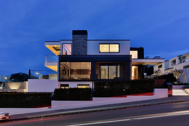 Twilight exterior view of a contemporary multi-level residence in an urban setting. The house has a flat-roofed composition, white rendered walls, dark vertical screening and slatted facade elements, extensive glazing, projecting balconies, and a prominent dark stone-clad chimney or feature wall. A landscaped frontage, boundary walls, steps, and street are visible.