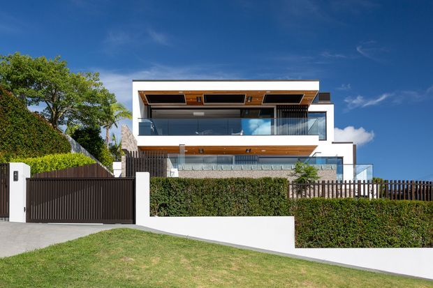 Front exterior view of a multi-level contemporary residence on a sloping site. The home has strong horizontal massing, crisp white rendered walls, extensive glass balustrades and glazing, warm timber-lined soffits, projecting balconies, and a flat roof. A dark vertical-slat driveway gate, rendered boundary walls, hedges, and landscaped planting define the street frontage.