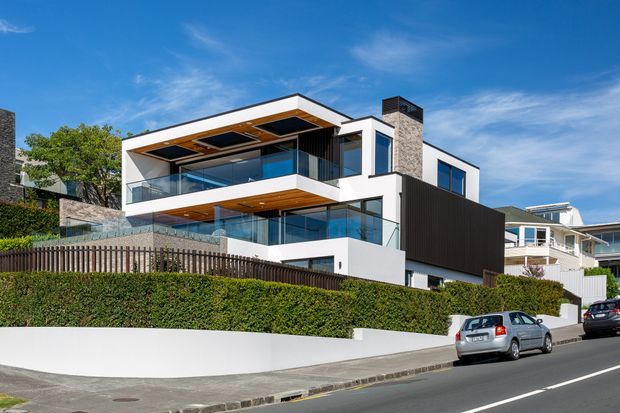 Street-level exterior view of a multi-storey contemporary residential home with crisp white rendered walls, dark vertical cladding, extensive glass balustrades and glazing, warm timber-lined soffits, and a flat projecting roof form. The property sits behind landscaped hedges and a low white boundary wall on a suburban street, with neighbouring homes visible.