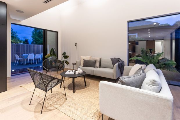 Bright contemporary living room with pale timber flooring, a large woven rug, two light upholstered sofas, black woven lounge chairs, and a round black coffee table. Full-height glazing opens views to a furnished outdoor patio and landscaped courtyard, while a dark built-in wall and recessed ceiling lighting add contrast.