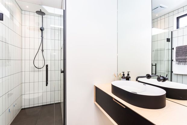 Bright contemporary bathroom featuring a frameless glass shower enclosure with white grid-pattern wall tiles, dark floor tiles, matte black shower fittings, and a floating timber-toned vanity with a sculptural black-and-white vessel basin. A large mirror reflects the vanity and a towel rail, while black tapware and accessories provide contrast.