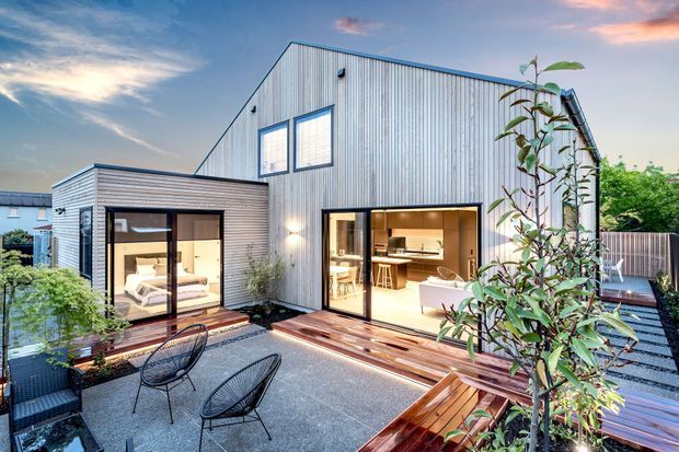 Exterior view of a contemporary multi-level home with corrugated metal cladding, extensive glazed sliding doors, and a rooftop or elevated timber deck courtyard. The image shows a bedroom through the left glazing and a warmly lit kitchen/living area through the central doors, with outdoor seating and planting in the foreground.
