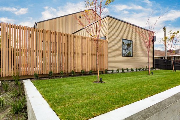 Exterior view of a contemporary low-rise building with horizontal light timber cladding, a taller dark-toned upper volume, black-framed window, and a tall vertical timber privacy fence. A neatly landscaped lawn, young trees, garden planting, and concrete retaining or edging walls surround the building.