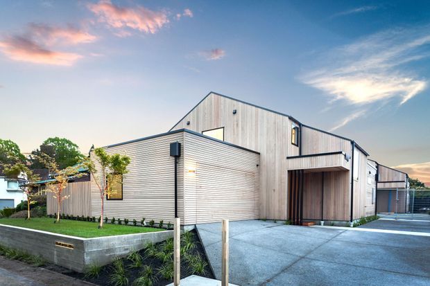 Exterior view of a contemporary multi-level building with pale vertical weatherboard cladding, dark metal roof edging and frames, and a large recessed timber-lined entry. The property includes a concrete driveway, landscaped raised garden beds, and a separate covered parking or service structure.