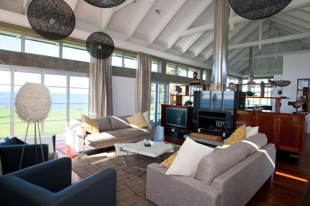 A bright, spacious living room with dramatic exposed white-painted roof trusses and high ceilings. Large floor-to-ceiling windows and clerestory glazing overlook a coastal landscape. The room contains two light-grey sofas, dark blue armchairs, a glass coffee table, a fireplace and media joinery, timber cabinetry, patterned rug, sheer curtains, and oversized woven pendant lights. The interior has a refined contemporary feel with strong natural light and timber accents.