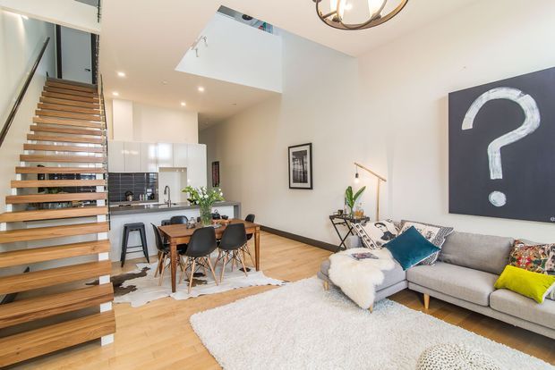 Bright open-plan contemporary interior showing a living room, dining area, and compact kitchen beneath a double-height void. A floating timber staircase with dark metal supports dominates the left side, while the living area has a large grey sectional sofa, pale rug, and statement artwork. Warm timber flooring contrasts with white walls and cabinetry, and an upper-level landing is visible above.
