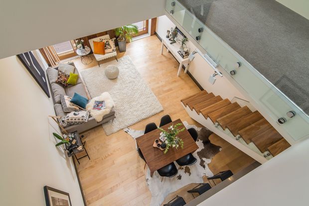 High-angle interior view of a stylish open-plan living and dining area. A grey sectional sofa, textured light rug, coffee table, plants and soft furnishings occupy the living zone, while a dark timber dining table with black chairs sits below a staircase. The staircase has warm timber treads, a pale structural side wall and a glass balustrade. Light timber flooring and white walls create a bright, contemporary atmosphere.