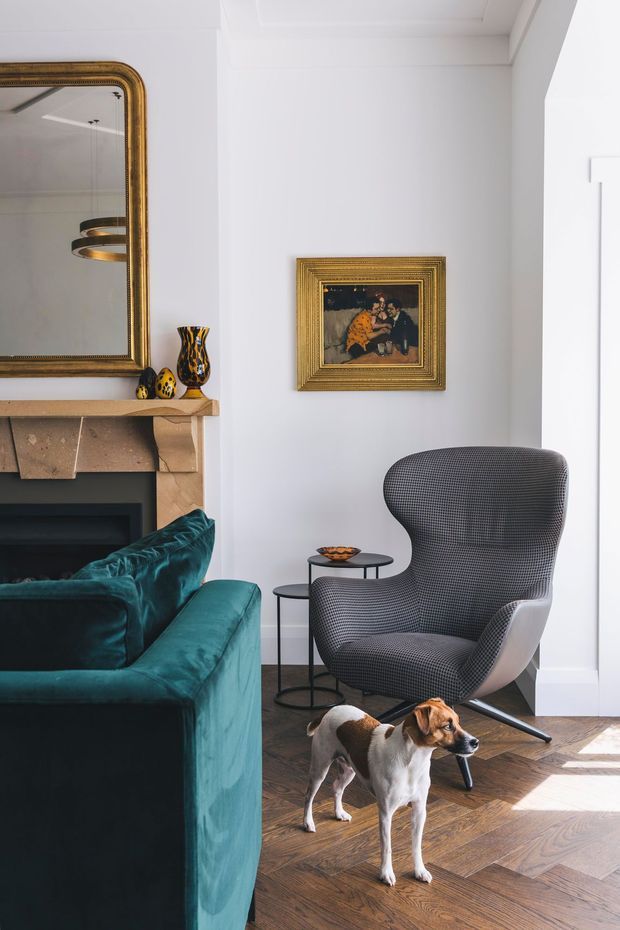 A refined living room corner with white walls and detailed ceiling trim, a deep teal velvet sofa, a sculptural patterned wingback lounge chair, and small round side tables. A carved stone or timber fireplace mantel with a large antique-style gold mirror and framed artwork adds traditional character, while herringbone timber flooring and modern lighting create a polished layered interior. A dog is present in the foreground.