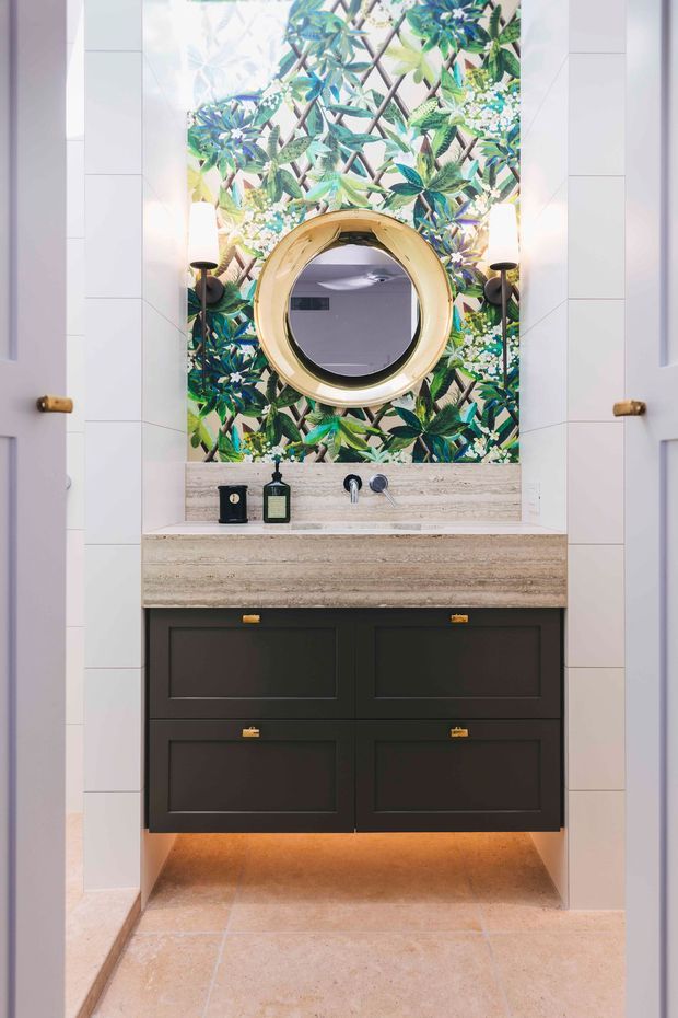 A compact, highly styled bathroom vanity alcove framed by open white doors. The space features a dark charcoal floating vanity with brass pulls, a pale travertine or stone benchtop and integrated backsplash, a round brass-framed mirror, wall sconces, and a vivid botanical mural behind the mirror. Pale stone or ceramic floor and wall tiles create a bright backdrop, with warm under-vanity lighting.
