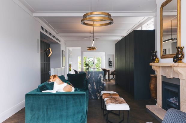 A refined open-plan living room with a deep teal velvet sofa in the foreground, a patterned armchair, and a pair of leather-and-fabric stools. The room features dark timber or black built-in cabinetry, a decorative pale stone or painted fireplace with a large gold-framed mirror, white coffered ceiling detailing, and layered circular pendant lighting. A dining area and bright garden outlook are visible beyond the living space. The overall scheme is elegant, high-contrast, and contemporary with classic detailing.