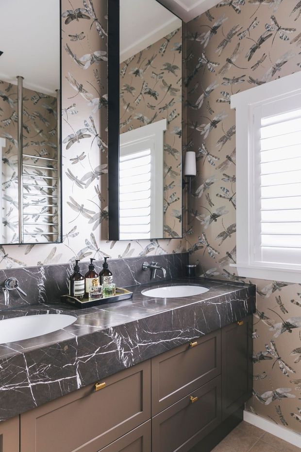 A refined bathroom with a double vanity, dark veined stone benchtop and matching splashback, taupe shaker-style cabinetry, patterned botanical wallpaper, black-framed mirrors, chrome tapware, and a white plantation-shuttered window. The space is bright and detailed, with a sophisticated traditional-meets-contemporary character.