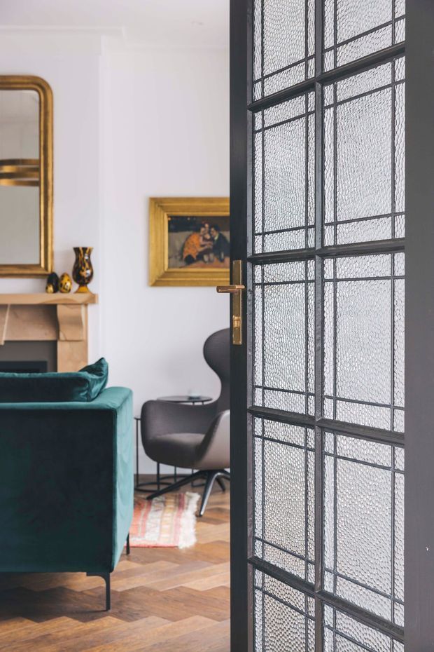Interior view into a refined living room through a partially open black-framed glazed door. The room features white walls, a classical fireplace with a gold-framed mirror and artwork, a deep teal velvet sofa, dark lounge chairs, a patterned rug, and warm herringbone timber flooring. The prominent door has a grid of textured translucent glass panels with dark metal framing and a brass-toned handle.