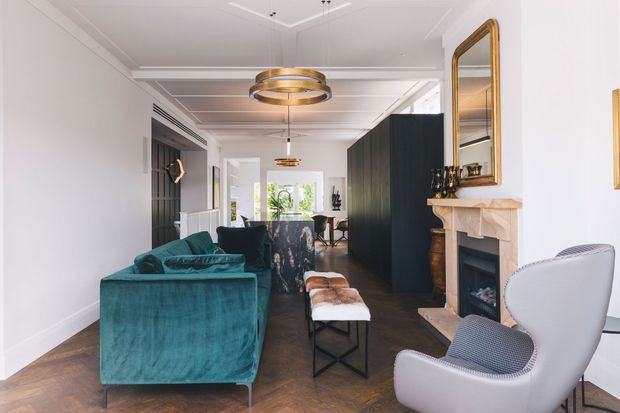 Refined open-plan living and dining room with a teal velvet sofa, sculptural lounge chairs, dark built-in cabinetry, a marble or stone fireplace surround, herringbone timber flooring, ornate wall mirror, and layered contemporary pendant lighting. The room combines classic architectural detailing such as decorative ceiling mouldings and a fireplace with modern furnishings and dark joinery.
