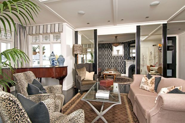 Elegant living room with a traditional fireplace, ornate black-and-white feature wallpaper, mirrored panels, dark timber floors, a glass coffee table, a blush-pink sofa, patterned armchairs, and a decorative sideboard. The room has strong symmetry, layered textiles, and refined period-inspired detailing.