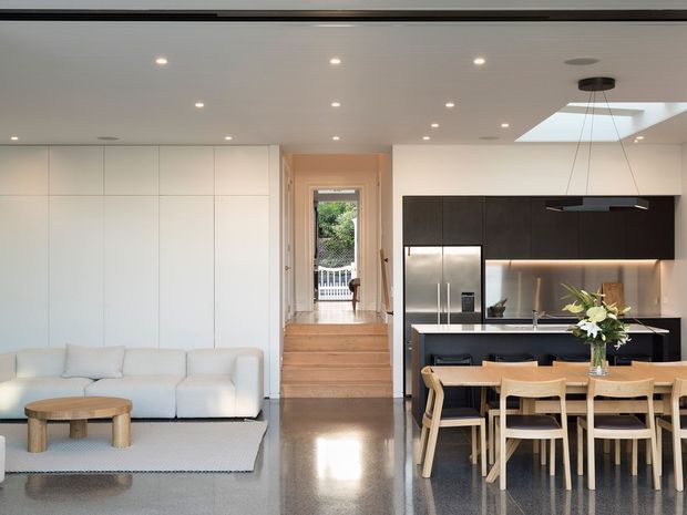 Open-plan contemporary interior combining a living room, dining area and kitchen. The space features a white built-in wall, pale sectional sofa, polished dark grey floor, timber dining furniture, dark kitchen cabinetry, stainless-steel appliances, a light benchtop, recessed ceiling lights and a skylight above the kitchen/dining zone. A short set of timber steps leads to a glazed rear doorway.