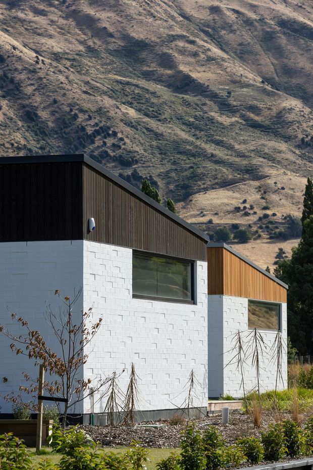 Exterior view of two contemporary gable-roofed buildings set against a steep, dry mountainous landscape. The buildings combine white painted masonry block lower walls with dark vertical timber-like cladding and black metal roof edges, large horizontal windows, and landscaped foreground planting.