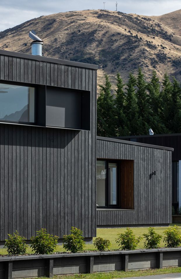 Exterior view of a contemporary dark-clad house composed of overlapping low, box-like volumes. Vertical charcoal cladding, large black-framed windows, timber detailing around an entry, and rooftop flues are visible. The home is set against a rugged dry mountain landscape with tall evergreen screening and a landscaped front edge.