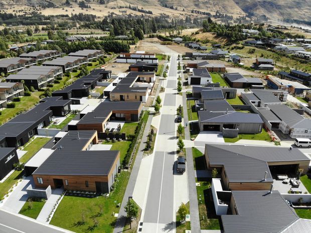 Aerial view of a contemporary suburban residential development arranged along a landscaped central street. Numerous detached, low-profile homes feature dark pitched roofs, rectangular forms, light-rendered walls, timber-toned cladding, private lawns, paved driveways and boundary fencing. Dry rolling hills and established vegetation form the background.