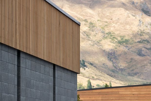 Close-up exterior architectural view showing a contemporary building facade composed of dark grey modular masonry or concrete panels below vertically ribbed timber cladding. A clean sloping roof edge and a secondary warm timber-clad structure are visible, with rugged mountain terrain in the background.