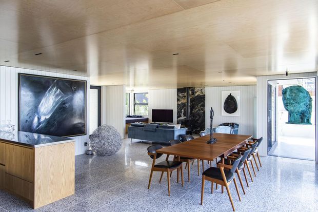 Open-plan contemporary living and dining area with a long timber dining table, mid-century-style chairs, a grey sofa, television, large artworks, pale timber ceiling, vertical wall panelling, and polished terrazzo flooring. Large glazed openings provide abundant natural light and connect to an outdoor area.