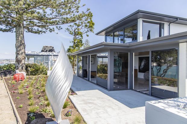 Bright exterior view of a contemporary two-storey residence with a flat roof, strong horizontal lines, white rendered walls, dark-framed floor-to-ceiling glazing, and a broad paved terrace or entry path. A sculptural white garden element and landscaped planting sit in the foreground, while water and distant urban scenery are visible beyond the property.