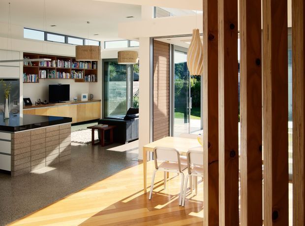 Bright open-plan contemporary home interior showing a kitchen, dining area and living room. The space features a tiled kitchen island, extensive timber cabinetry and shelving, polished concrete flooring in the living area, warm timber flooring in the dining zone, large glazed doors and windows, and a high ceiling with clerestory glazing. Vertical timber screening partially frames the foreground.