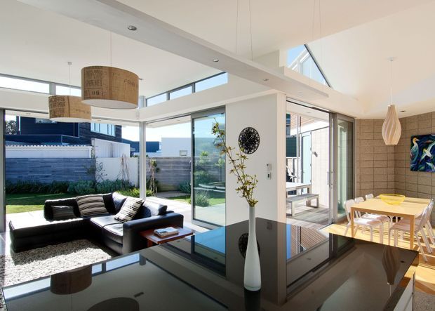 Bright, open-plan contemporary living and dining area with extensive floor-to-ceiling glazing opening to a landscaped courtyard. A black sectional sofa sits on a textured rug beside a glossy dark reflective coffee table, while a light timber dining table and chairs occupy the adjacent zone. White walls and ceilings, exposed timber decking outside, a tiled feature wall in the dining area, sculptural pendant lights, and a roof skylight create an airy indoor-outdoor connection.