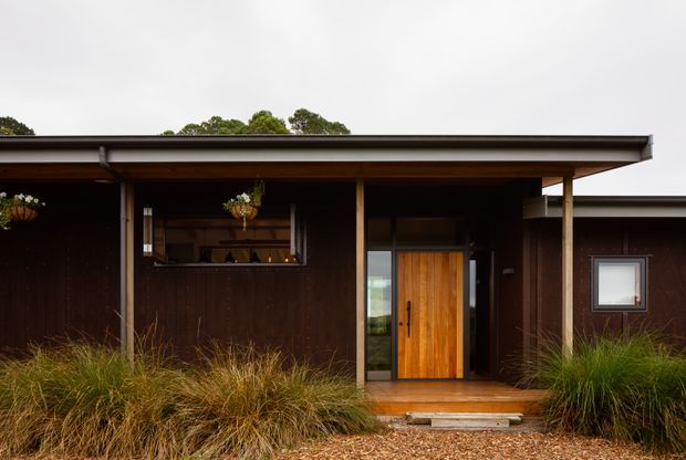 Single-storey dark timber-clad house facade with a broad low-pitched roof, covered entry porch, warm timber front door, and a small serving or pass-through window. Ornamental grasses and gravel landscaping frame the entrance. The image focuses on the residential exterior.