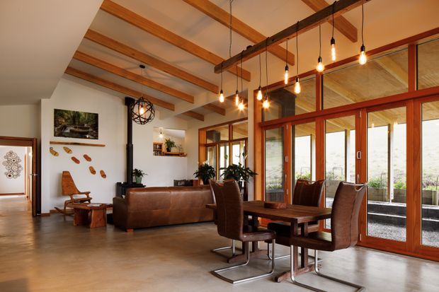 Warm, open-plan living and dining area with exposed timber ceiling beams, polished concrete flooring, a dark wood-burning stove, leather seating, and a substantial timber dining table. Full-height timber-framed glazed doors and windows line one side, bringing in abundant natural light. Suspended pendant lights and timber joinery create a rustic yet refined, lodge-like atmosphere.