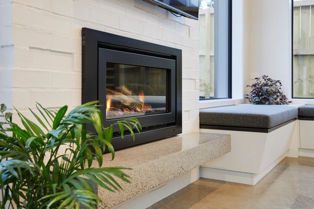 Bright contemporary living area featuring a built-in black-framed fireplace set into a painted white brick wall, with a substantial terrazzo hearth extending toward a white built-in window seat. Large windows provide natural light, while indoor plants add greenery. The space has polished concrete or stone-like flooring and a clean, refined aesthetic.
