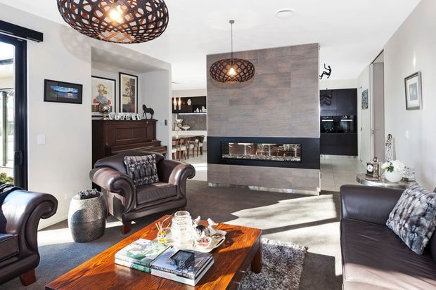 Bright contemporary living room with a feature fireplace wall clad in grey-brown timber-look panels, a long inset firebox, dark leather seating, and a substantial polished timber coffee table. A dining area and kitchen are visible beyond the open-plan space. Woven pendant lights, artwork, patterned cushions, and layered rugs add warmth and texture.