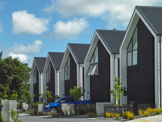 Exterior view of a row of closely spaced contemporary gable-front townhouses. The buildings feature steep dark corrugated metal roofs, charcoal vertical cladding, light grey structural frames and white horizontal side cladding, with large vertical windows and landscaped front boundaries. Several blue vehicles are parked along the street, with trees and a bright partly cloudy sky in the background.
