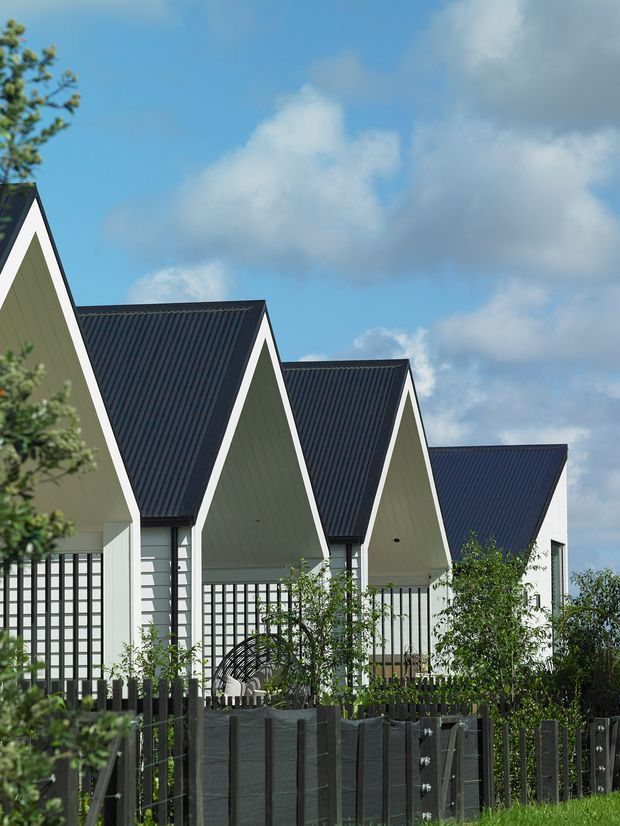 Exterior view of a row of closely arranged gabled buildings with steep dark corrugated metal roofs, pale green vertical cladding, crisp white trim, and extensive gridded glazing. A dark timber privacy fence and landscaped planting occupy the foreground beneath a bright partly cloudy sky.