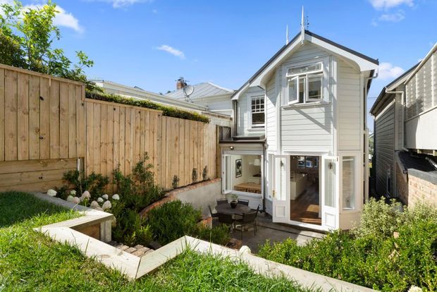 Bright exterior view of a narrow, multi-level residential dwelling with white horizontal weatherboard cladding, a pitched gable roof, projecting bay window, and glazed French doors opening to a small paved courtyard. Timber privacy fencing, raised garden beds, lawn, and neighbouring houses are visible.