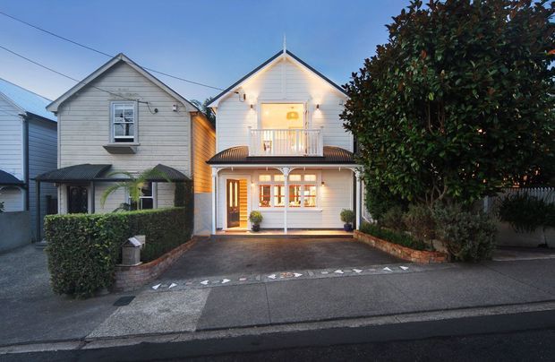 Street-facing view of a compact two-storey detached home with a symmetrical gabled roof, white weatherboard cladding, a covered balcony, illuminated ground-floor entry, and a small paved driveway bordered by hedges and brick edging. The adjacent homes and mature tree are visible in the residential streetscape.