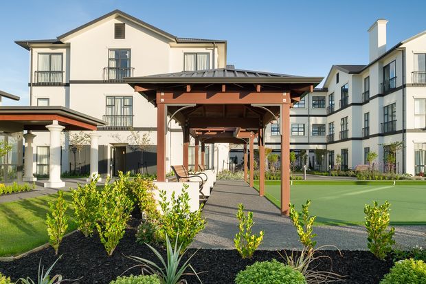 Exterior view of a multi-storey residential complex with pale rendered facades, dark pitched roofs, balconies, and black-framed windows. A timber-covered pedestrian pergola runs through landscaped communal grounds, with paved paths, lawn, planting beds, and a covered outdoor seating area.