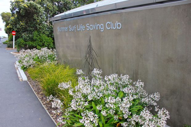Exterior view of the Summer Surf Life Saving Club, featuring a simple rendered concrete or masonry wall with raised lettering and a subtle line-art graphic. A planted garden bed with grasses and flowering shrubs runs along the wall beside a paved pedestrian path and road signage.