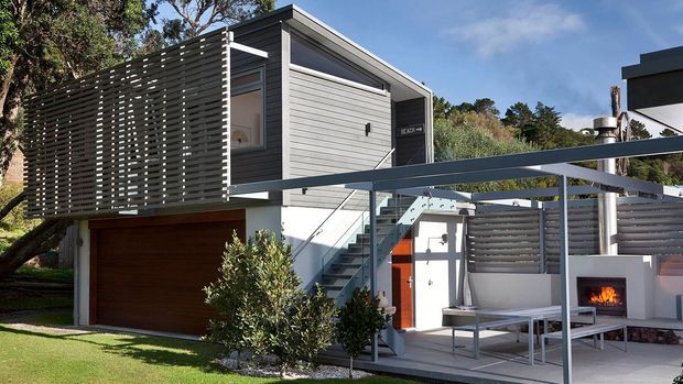 Exterior view of a contemporary multi-level home with a raised upper volume clad in horizontal grey weatherboards, a timber garage door, external metal stairway, slatted privacy screening, and a covered outdoor entertaining area with built-in fireplace. The design uses clean geometric lines and a restrained grey-and-white palette integrated into a landscaped suburban setting.