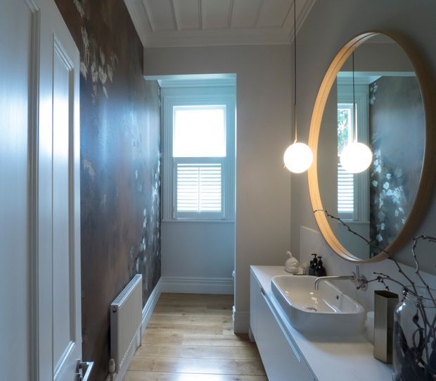 A narrow, refined bathroom with a white vanity and vessel sink, large oval wood-framed mirror, pendant globe lighting, pale timber flooring, decorative dark botanical wall finish, white trim, and a bright shuttered window. A hallway-like passage leads toward the window, with a radiator along the feature wall.