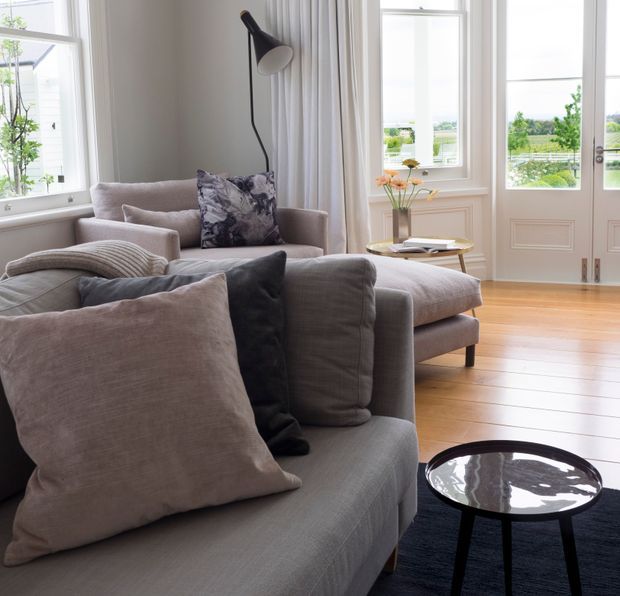 A bright, refined living room with large windows and glazed double doors overlooking landscaped grounds. The room features layered grey upholstered sofas, a chaise, patterned and textured cushions, a round black glass coffee table, a sculptural black floor lamp, sheer curtains, a small side table, and warm timber floorboards. Decorative wall panelling and tall windows add a classic architectural character.