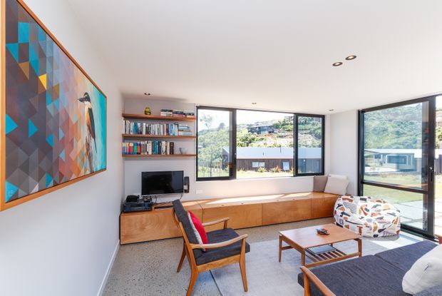 Bright mid-century-inspired living room with a large colourful geometric artwork, timber built-in media and window-seat cabinetry, open shelving, a television, mid-century lounge chairs, a coffee table, and extensive black-framed glazing overlooking a green suburban setting. The room has a polished speckled concrete or terrazzo-like floor and a clean white ceiling with recessed downlights.